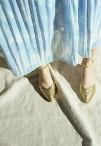 Goldene spitze Schuhe mit einem strukturierten Muster und Knöchelriemen, kombiniert mit einem hellblauen, plissierten Rock, vor einem neutralen Hintergrund.