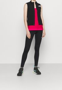 Černá bezrukávová vesta, jasně růžový top, černé legíny a černé běžecké boty s zelenými akcenty. Jednoduchý design, přiléhavý styl.