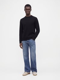 Mand stående iført en sort langærmet sweater, lyseblå vidde-ben jeans og beige sneakers mod en ensfarvet hvid baggrund.