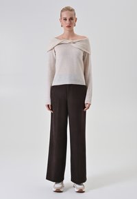 Maglione lungo color avorio con scollo ampio e drappeggiato, abbinato a pantaloni scuri a gamba larga e scarpe da ginnastica bianche con accenti marroni.