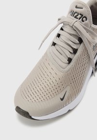 Nike Air Max 270 sportcipő világos bézs hálós anyagból, texturált felsőrésszel, lapos cipőfűzőkkel, fekete részletekkel és fehér gumitalppal.