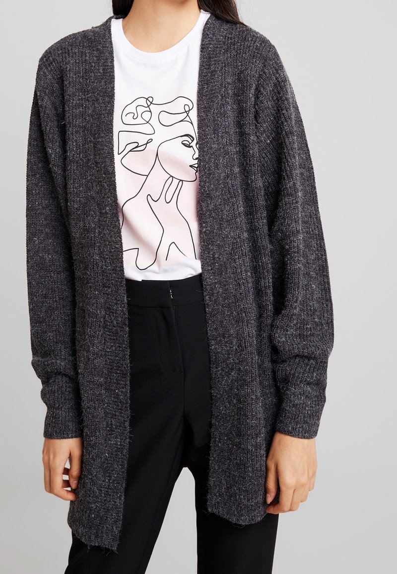 Dunkelgrauer Strickcardigan mit offenem Frontbereich, langen Ärmeln und strukturiertem Stoff. Getragen über einem weißen Shirt mit einem minimalistischen Linienstil-Design.