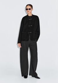 Vrouw die een zwarte oversized jas met sluitingen met knopjes draagt, wijde broek, zwarte puntige schoenen en rechthoekige zwarte zonnebril tegen een effen achtergrond.