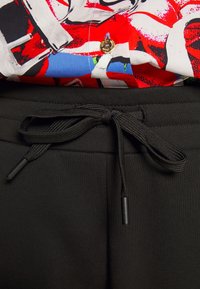 Černé tepláky mají strukturovanou tkaninu, pas se stahovací šňůrkou s kovovými koncovkami a jsou spárovány s vícebarevným vzorovaným tričkem.