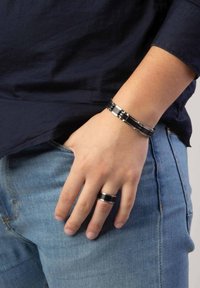 Zwarte en zilveren metalen armband met elkaar vergrendelde schakels, gedragen om de pols, naast een zwarte bandring. Casual spijkerbroek zichtbaar.