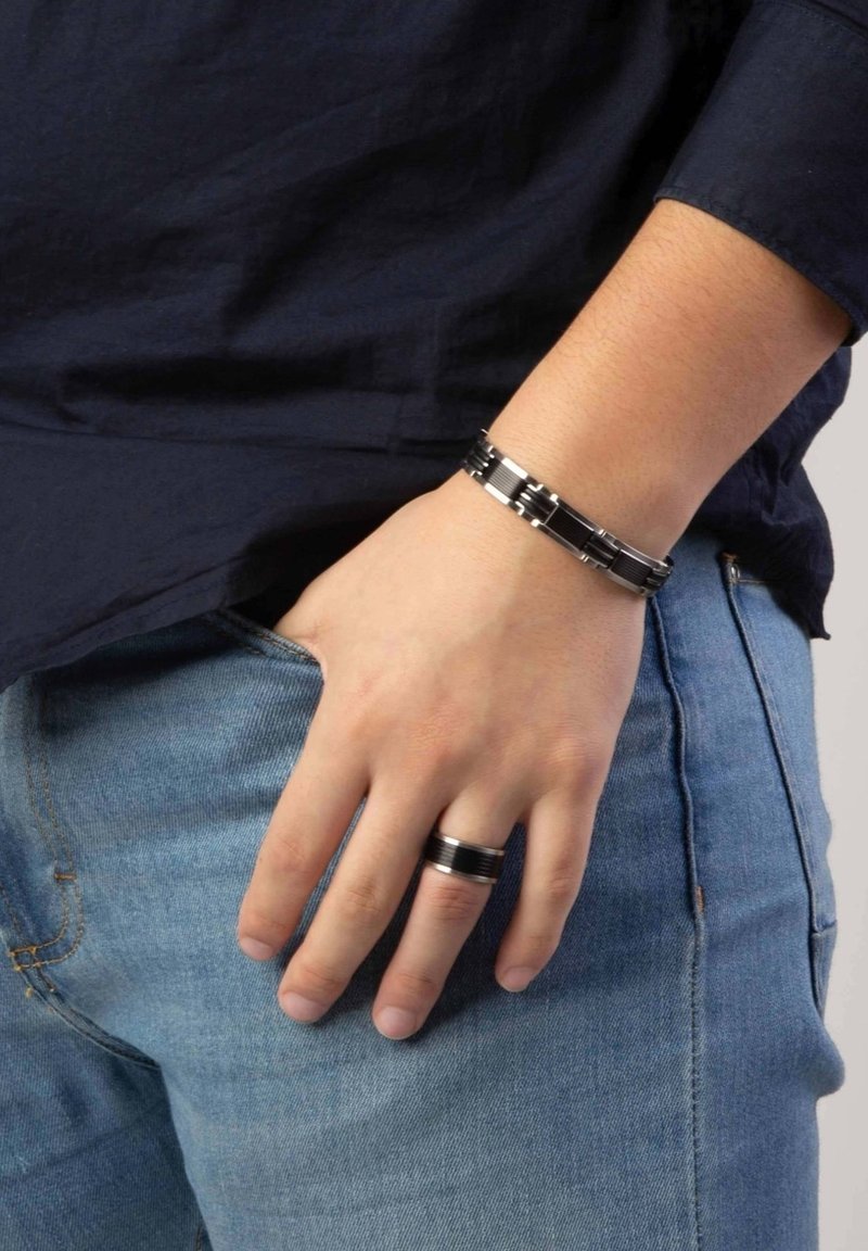 Zwarte en zilveren metalen armband met elkaar vergrendelde schakels, gedragen om de pols, naast een zwarte bandring. Casual spijkerbroek zichtbaar.