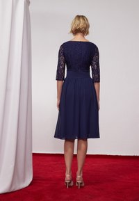 Robe bleu marine avec des manches en dentelle trois-quarts, corsage ajusté avec une taille froncée et jupe fluide, assortie à des talons de couleur claire.