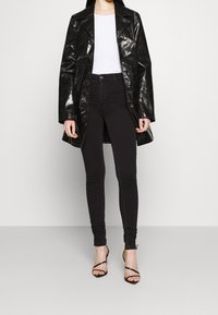 Trench coat negro brillante con un cuello puntiagudo, combinado con jeans negros de cintura alta y tacones negros con tiras. Top blanco sencillo debajo.