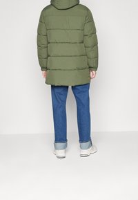 Person iført en grøn quilted pufferjakke, blå manchetjeans og hvide sneakers, stående foran en ensfarvet lys baggrund.