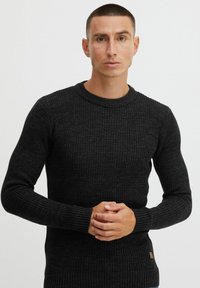 Black ribbed knit sweater with long sleeves and round neckline. Textured fabric, fitted design, featuring a small logo patch at the hip.