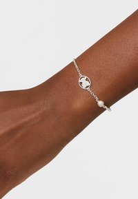 Braccialetto in argento con un piccolo ciondolo a forma di farfalla rotonda e una singola perla bianca indossato su un polso scuro su uno sfondo chiaro.
