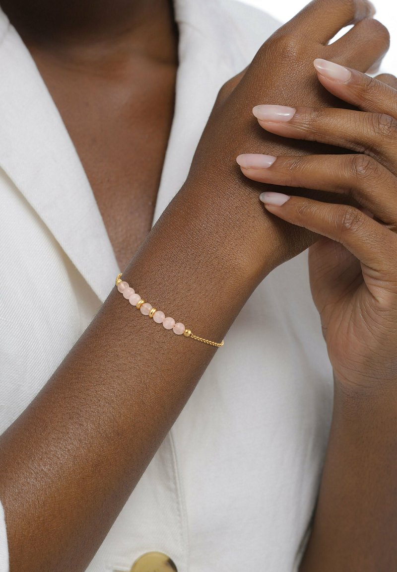 Bracelet en chaîne dorée orné de perles roses à la texture lisse, agrémenté de petits disques en or, porté sur une peau de ton brun.