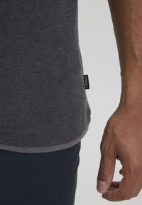 Czarna krótka koszulka w odcieniu ciemnoszarym, wykonana z miękkiego materiału, z zaokrąglonym dołem i czarną metką z białym tekstem na dolnej stronie.