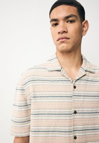 Camicia a maniche corte color beige chiaro con motivi a righe orizzontali blu e bianche, dotata di colletto a punta e bottoni scuri.