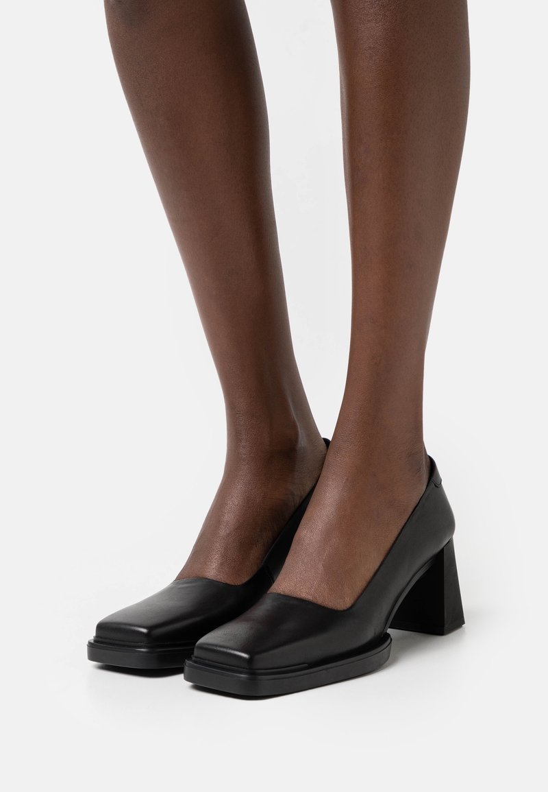 Jambes à la peau foncée portant des chaussures à talons carrés en cuir noir, debout sur une surface blanche.