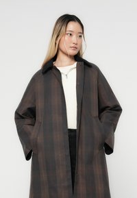 Lang, mørkternet frakke med knaplukning, sidelommer og krave. Har et blødt, tekstureret stof med nuancer af brun og grå.