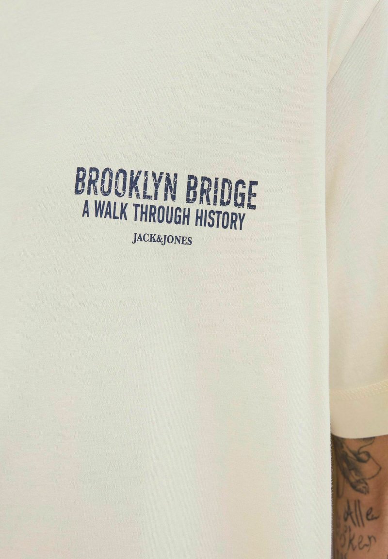 Pamutból készült krémszínű póló, amelyen sötétkék felirat olvasható: "BROOKLYN HÍD EGY SÉTA A TÖRTÉNELEMEN KERESZTÜL JACK&JONES." Rövid ujjú, egyszerű dizájn.
