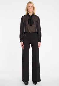 Zwarte doorschijnende blouse met een strik bij de halslijn, aangevuld met zwarte wijde broek met subtiele sprankels.