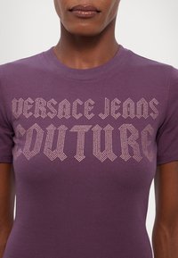 Fialové tričko s krátkým rukávem z měkké bavlny, se zdobením z kamínků "Versace Jeans Couture" přes hruď.