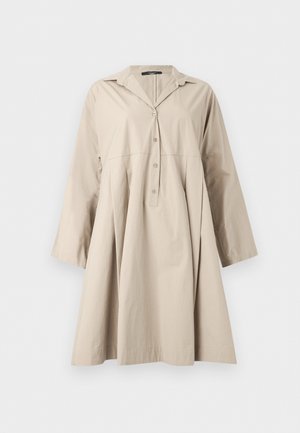 Femme debout portant une robe beige boutonnée mi-genou avec manches longues et des chaussures plates marron foncé avec brides à la cheville.