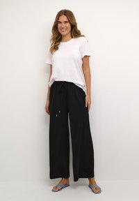 Witte katoenen t-shirt gecombineerd met losse zwarte broek met een elastische tailleband en trekkoord. Blauwe slippers maken de outfit compleet.