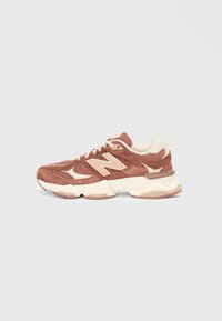 Baskets en suédine marron avec des accents crème, présentant un design texturé, un système de laçage et une semelle rembourrée. Logo classique de New Balance visible.