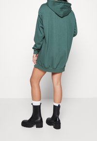 Sweat à capuche vert oversize avec capuche à cordon, texture douce et poignets côtelés, associé à des bottines noires épaisses et des chaussettes blanches.
