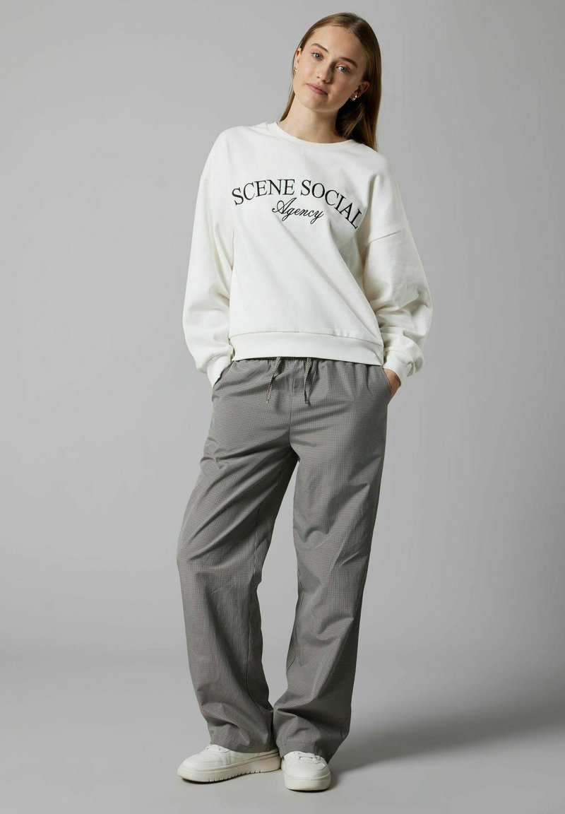 Sudadera blanca con el texto "Agencia SCENE SOCIAL", combinada con pantalones sueltos de cuadros grises y zapatillas blancas.