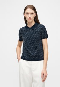 Γυναίκα που φοράει μπλε κοντομάνικο πόλο με λευκές πουά γραμμές και λευκό ψηλόμεσο παντελόνι, στέκεται με το ένα χέρι στην τσέπη.