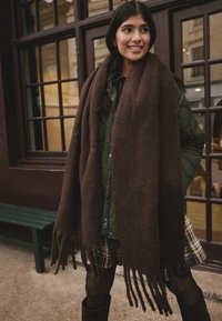 Brown, knitted wool scarf with fringes, wraps around the shoulders, paired with a green quilted jacket and a plaid skirt.