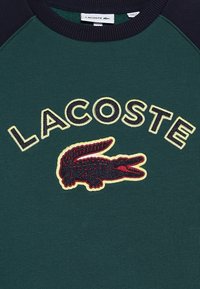 Maglione verde con maniche raglan blu navy, caratterizzato da un logo Lacoste ricamato e un coccodrillo con accenti rossi e gialli. Tessuto morbido.