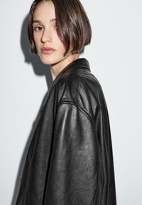 Zwarte leren jas met een gladde textuur en een ontspannen pasvorm, voorzien van een kraag met inkeping en opvallende stiksel-details.