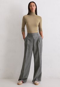 Frau steht gegen eine weiße Wand, trägt einen beigen Rollkragenpullover, hoch taillierte graue weite Hosen und beigen Sneakers mit roten Socken.