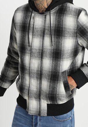 Mężczyzna ubrany w czarno-białą, kraciastą kurtkę na zamek z czarnymi mankietami i kapturem, z ręką w kieszeni, w zestawieniu z niebieskimi dżinsami.