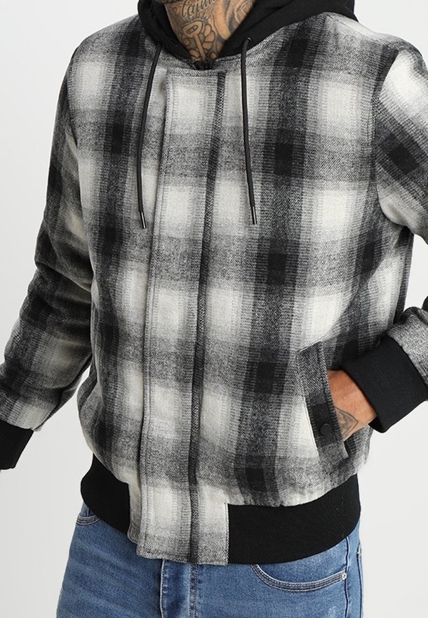 Homme portant une veste zippée à carreaux noir et blanc avec poignets noirs et capuche, main dans la poche, associée à un jean bleu.