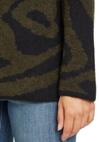 Main reposant sur le côté, portant un pull texturé à motifs noirs et verts olive et un jean en denim bleu clair.