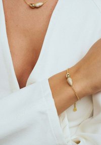 Guldring med en texturerad, oval berlock och små runda accenter. Bärs på ett handled, ihop med ett vitt plagg.