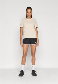 Μπεζ αθλητική μπλούζα με κοντά μανίκια σε συνδυασμό με μαύρο σορτς με λευκές λεπτομέρειες. Το μοντέλο φοράει μαύρα αθλητικά παπούτσια και λευκές κάλτσες.