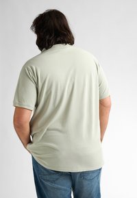 Ljusgrön kortärmad polo med en avslappnad passform, mjuk yta och klassisk krage. Buren med blå jeans, visad bakifrån.