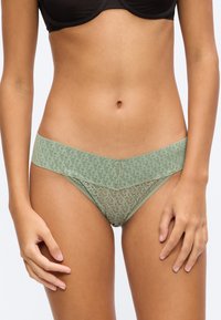 Donna che indossa biancheria intima in pizzo verde chiaro con motivi e un reggiseno nero, mostrata dal ventre alla parte superiore delle cosce, in piedi contro uno sfondo semplice.