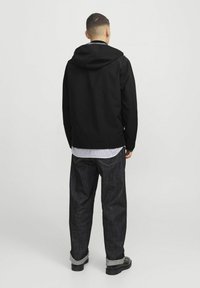 Giacca leggera nera con cappuccio, abbinata a jeans in denim blu. I jeans hanno un orlo arrotolato e sono indossati con scarpe nere.