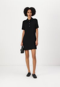 Abito polo nero con maniche corte, tre bottoni e orlo plissettato. Il materiale sembra liscio; la modella tiene in mano una piccola borsa nera.