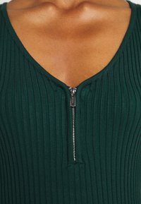 Rippierter grüner Bodysuit mit V-Ausschnitt und einem Reißverschluss auf der Vorderseite. Glatte Textur mit einem figurbetonten Design, das die Reißverschlussdetails und den Ausschnitt hervorhebt.