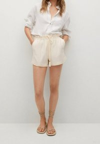 Chemise blanche à boutons, shorts crème avec cordon de serrage et poches, sandales plates avec liens à la cheville. Matière légère, coupe décontractée, style casual.