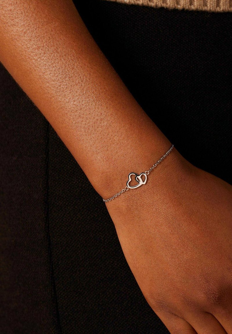 Zilveren kettingarmband met twee in elkaar vervlochten hartvormen. De armband ligt op een gestructureerd huidoppervlak.