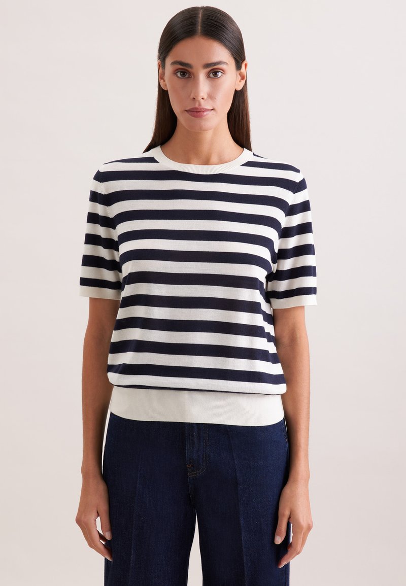 Femme aux cheveux raides et foncés portant un pull à manches courtes rayé blanc et bleu marine et un pantalon bleu foncé sur un fond uni.