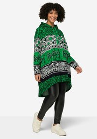Felpa verde a fantasia con dettagli neri e bianchi, dotata di cappuccio con laccetti e orlo asimmetrico, abbinata a leggings neri e sneakers bianche.