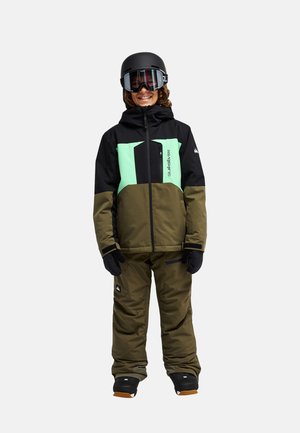 Enfant en veste et pantalon de neige verts et noirs, portant des gants noirs, un casque et des lunettes de ski, souriant et debout sur un fond uni.