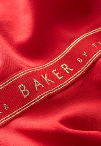 Rødt stof med en glat tekstur har en guldbroderet label, der lyder "BAKER by T." Labelen er rektangulær med en tynd kant.