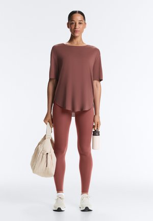 Kobieta stojąca w dopasowanym, pudrowo-różowym stroju sportowym, trzymająca beżową torbę na siłownię w jednej ręce i białą butelkę z wodą w drugiej.